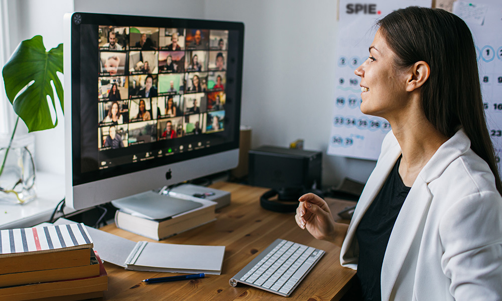 Female in white blazer sits in front of computer screen showing an online webinar setting with multiple faces